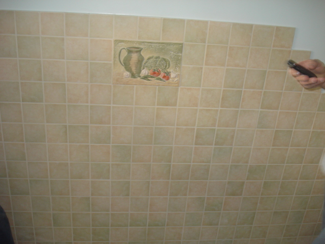 Ściana wyłożona jasnymi, kwadratowymi płytkami ceramicznymi z dekoracyjnym panelem przedstawiającym dzban i warzywa, przy której widać dłoń z nożem do cięcia płytek.